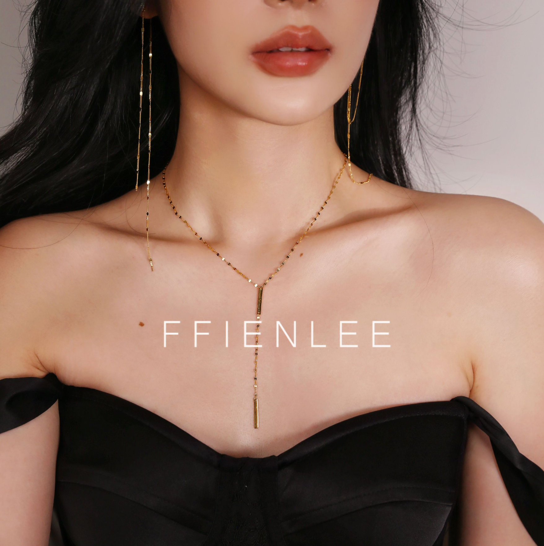 Close-up of model's chest and décolletage wearing FFIENLEE 925 sterling silver Y-lariat tile chain necklace with double bar pendant in 24K gold tone, Y-drop falling between the collarbones, paired with long gold tassel earrings, styled with a black off-shoulder dress, FFIENLEE branding